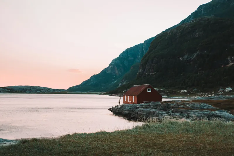 Lite rødt trehus på svabergene ved sjøen, med bratte fjell bak og rosa himmel ved soloppgang.