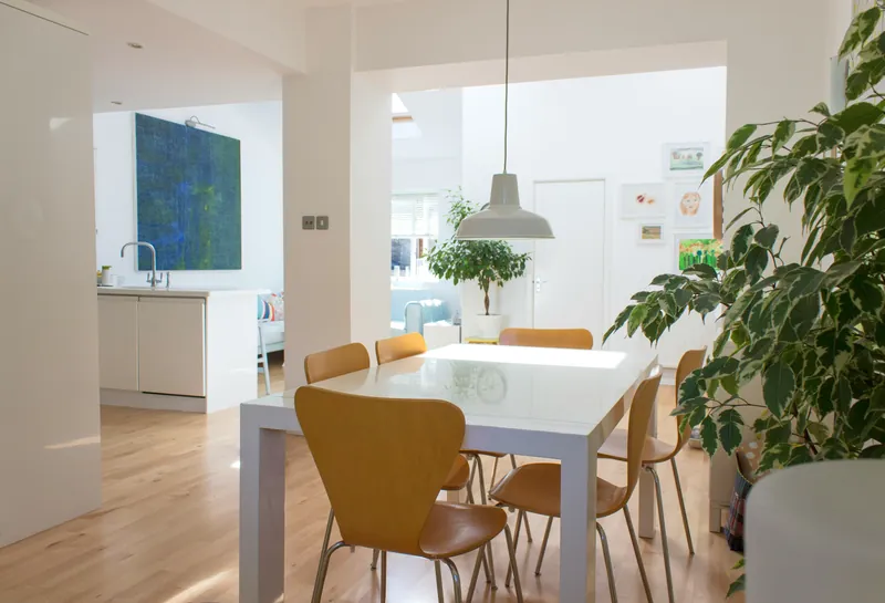 Lyst spiseområde med hvitt bord og stoler i lyst tre, en stor grønn plante og åpent kjøkken i bakgrunnen.